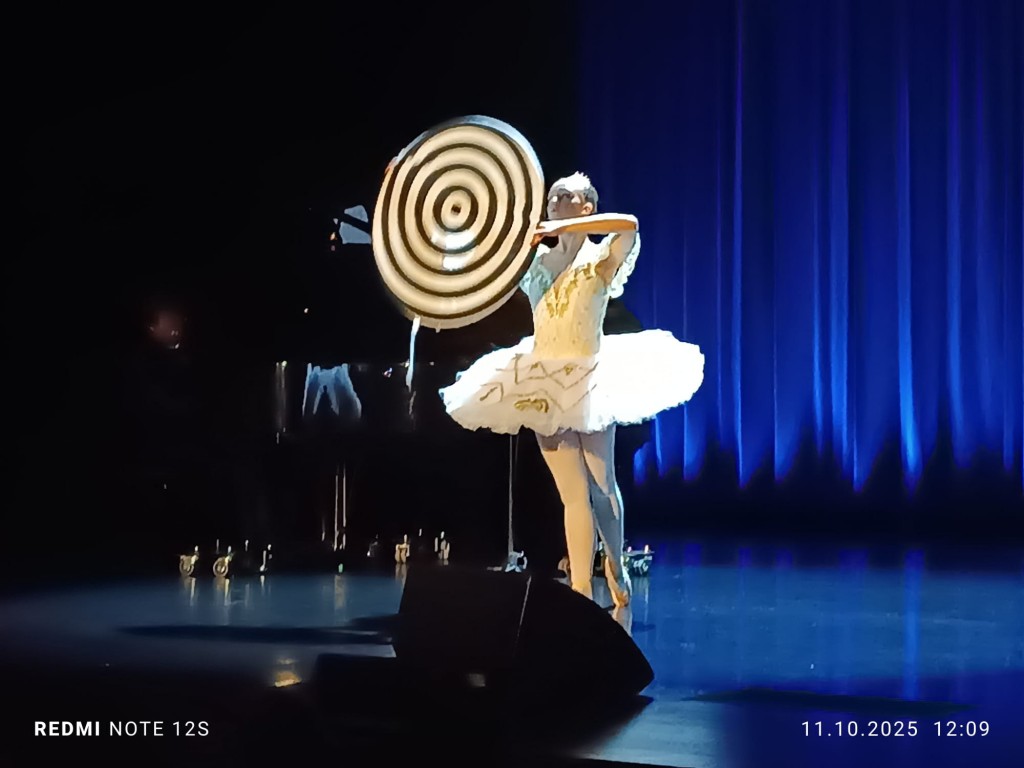 Widok na scene z prawej strony. Baletnica stoi na palcach, z uniesioną przed soba czarno-białą przypominającą spirale parasolką , w klasycznym, białym kostiumie scenicznym i tiarze. Za baletnicą widać pianistę grającego przy fortepianie, a całość oświetlo