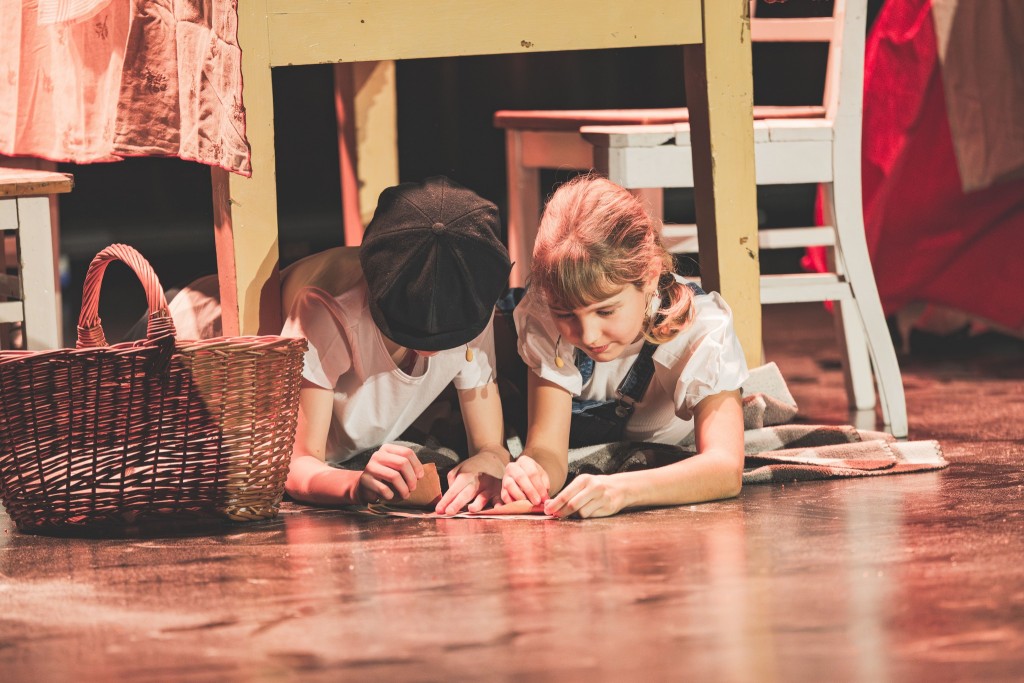 woje dzieci leży na podłodze pod stołem, skupione na wspólnym składaniu kartki. Jedno z nich ma na głowie ciemną czapkę z daszkiem, a drugie jasne włosy związane w kucyk. Obok nich stoi wiklinowy kosz, a cała scena jest oświetlona ciepłym, teatralnym świa