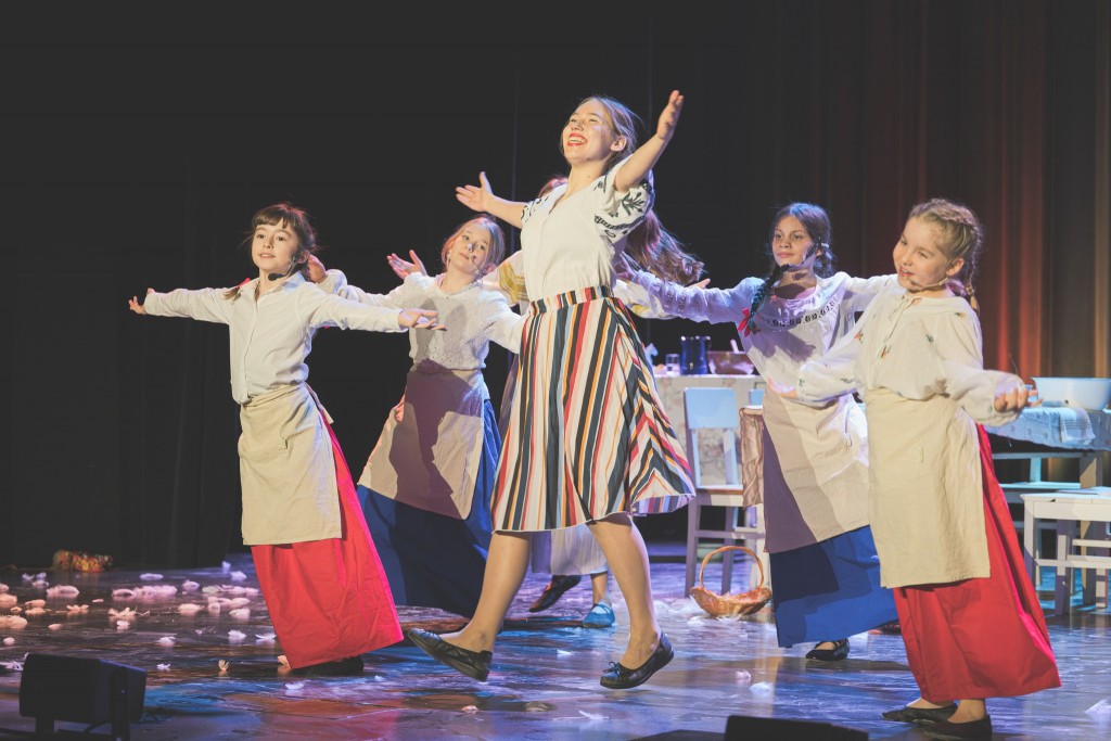 Widok z prawej strony. W centrum stoi pani w kolorowej, pasiastej spódnicy i białej bluzce, unosząca ręce z szerokim uśmiechem. Pozostałe dzieci mają na sobie długie spódnice i fartuchy w stylu ludowym i również wykonują taneczne gesty z rozłożonymi ramio