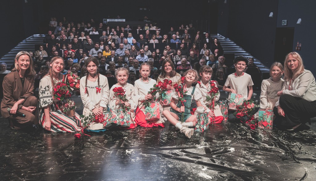 Widok z samej sceny. Dzieci siedzą na scenie uśmiechnięte z kwiatami i torbami prezentowymi z tyłu nich są rodzice i najbliżsi ,po prawej stronie zdjęcia kuca jedna pani a po lewej stronie zdjęcia pani dyrektor i pani nauczycielka teatru dla dzieci także 