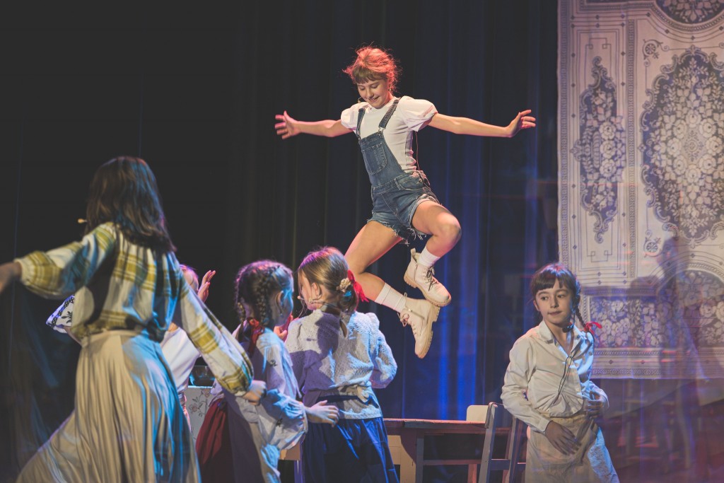 W centrum kadru jedna z dziewczynek skacze ze stolika wysoko w powietrze z szeroko rozłożonymi ramionami, ubrana w krótkie ogrodniczki i białe buty. Pozostałe dzieci, ubrane w różnorodne kostiumy, stoją poniżej i patrzą w jej stronę, biorąc udział w wspól