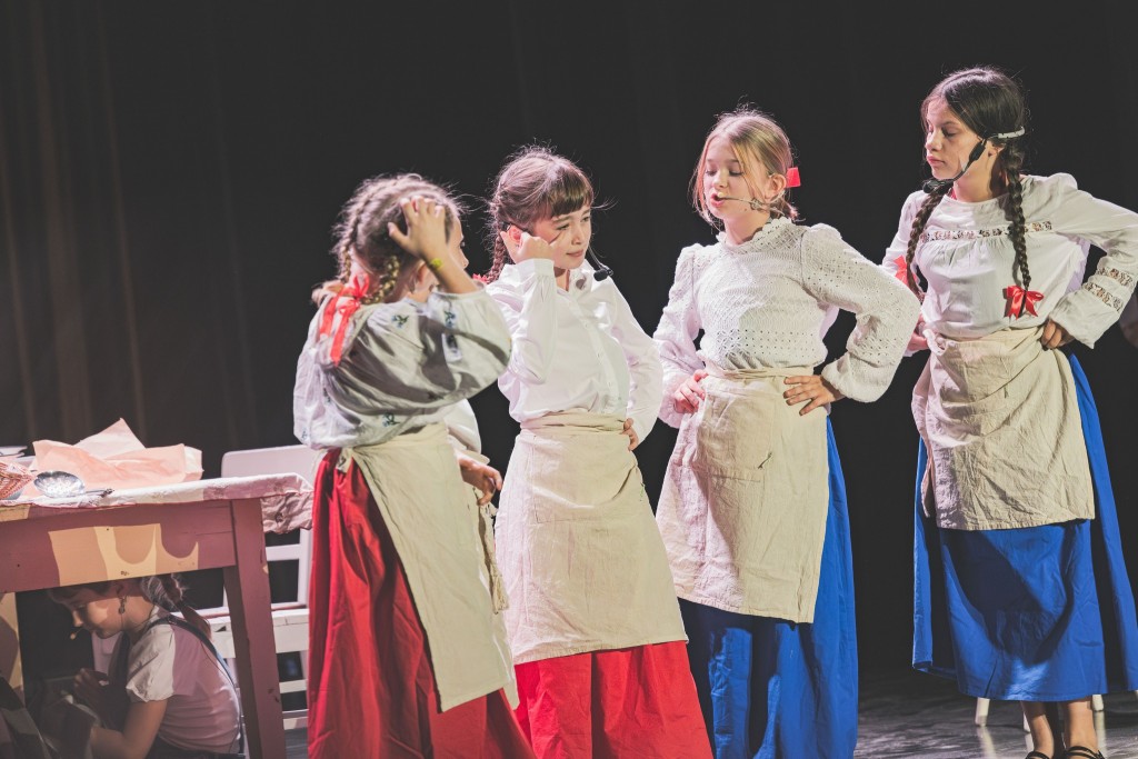 Na scenie stoi grupa dziewczynek w teatralnych kostiumach mają na sobie białe bluzki, długie spódnice w czerwieni lub błękicie oraz jasne fartuchy. Kilka z nich trzyma ręce na biodrach i wygląda, jakby prowadziły poważną rozmowę w ramach przedstawienia. W