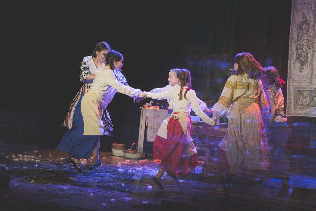 Na scenie kilka dziewczynek w strojach ludowych trzyma się za ręce i wiruje w kręgu, tworząc radosną scenę taneczną. Ubrane są w długie, kolorowe spódnice, białe bluzki z haftami oraz jasne fartuchy. W tle widać stół z rekwizytami kuchennymi i ozdobną tka