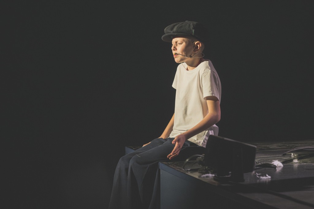 Widok z prawej strony. Na skraju sceny siedzi oświetlony chłopiec ma na sobie czarny berecik białą koszulkę i czarne spodnie .Mówi coś bardzo przejęty wykonując gest ręką. Wokół niego jest ciemno. Na scenie są ślady mąki i piórek.