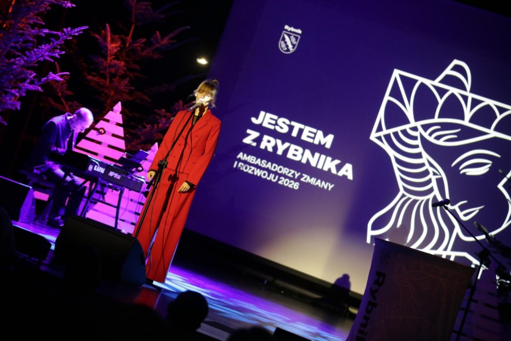 Sytuacja sceniczna. Kobieta w czerwonym garniturze śpiewa, w tle akompaniator przy instrumencie klawiszowym. Na ekranie Jestem z Rybnika, grafika i logo miasta.