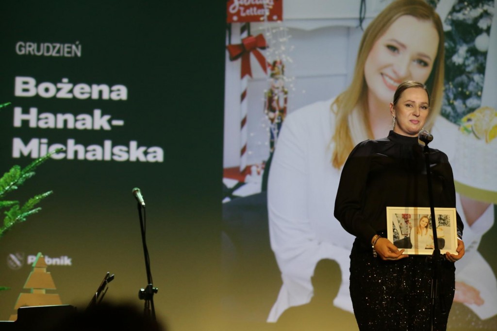 Kobieta na scenie przed mikrofonem, w rękach trzyma ramkę ze zdjęciem. W tle na ekranie zdjęcie tej samej kobiety.