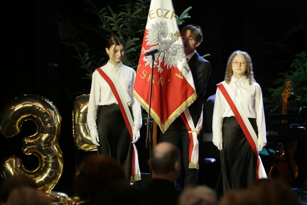 podczas jubileuszu, 3 osoby w biało czerwonych szarfach. Osoba w środku trzyma sztandar.
