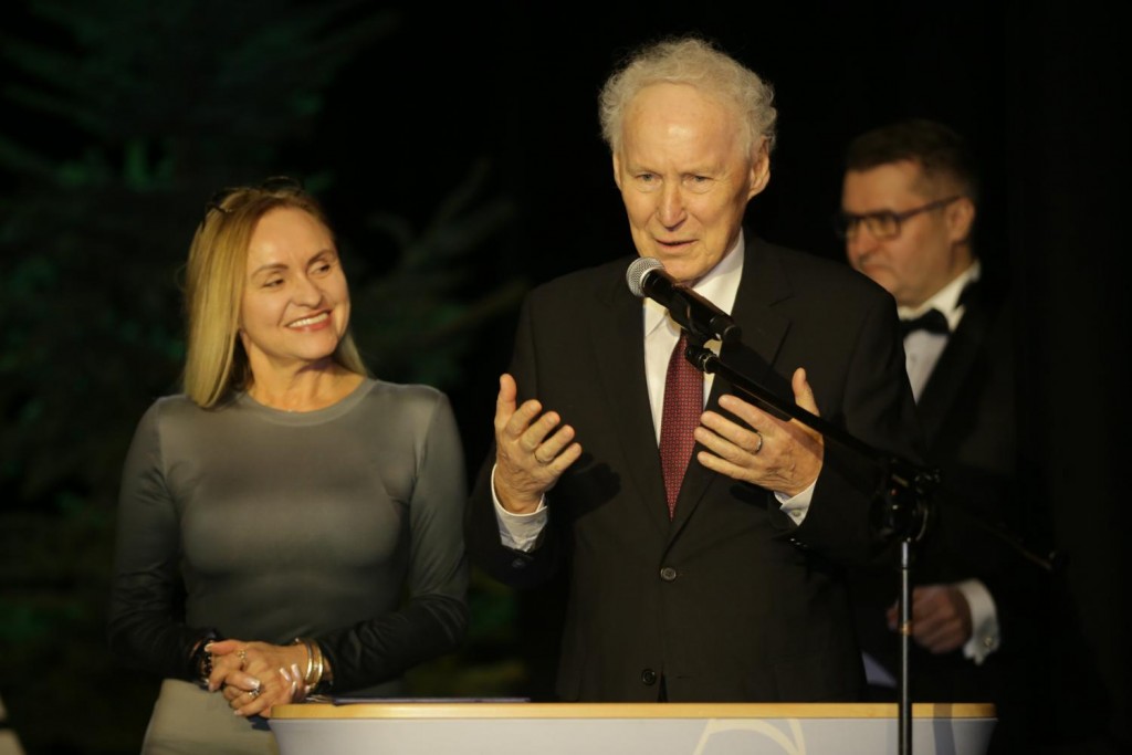 starszy mężczyzna przemawia, obok uśmiechnięta kobieta, w tle mężczyzna