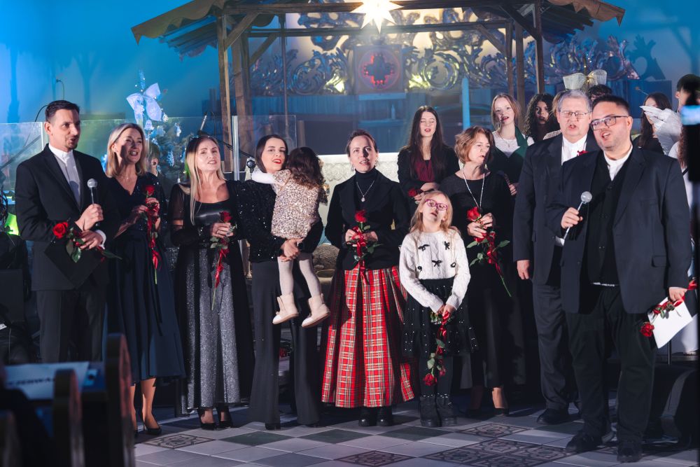 Sytuacja sceniczna, grupa osób śpiewa, część z nich trzyma w rękach róże. W tle stajenka.