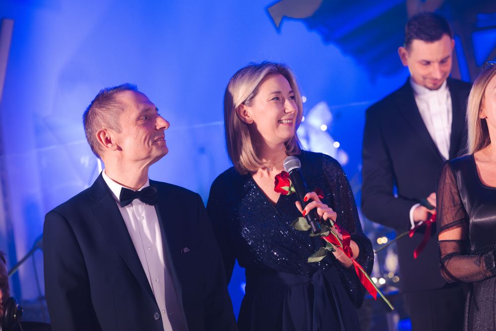 Dwoje odświętnie ubranych ludzi uśmiecha się i patrzy w bok, kobieta trzyma w ręce różę i mikrofon.