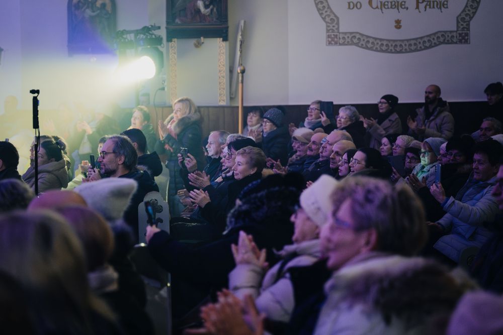 Publiczność siedzi w ławkach kościelnych i klaszcze. Widok z boku.