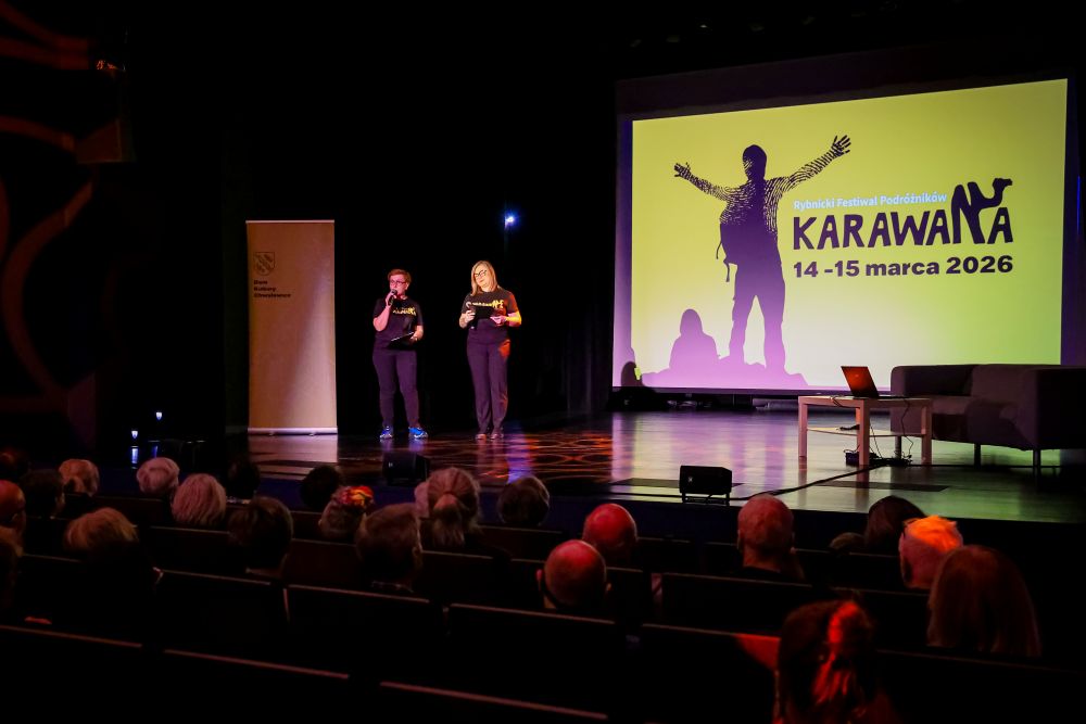 Dwie kobiety stoją na scenie. Jedna mówi do mikrofonu, druga - spogląda w notatki, oprócz nich na scenie znajduje się kanapa, a przed nią mały stolik z laptopem. W tle na rzutniku wyświetlana jest grafika “Karawana”. 