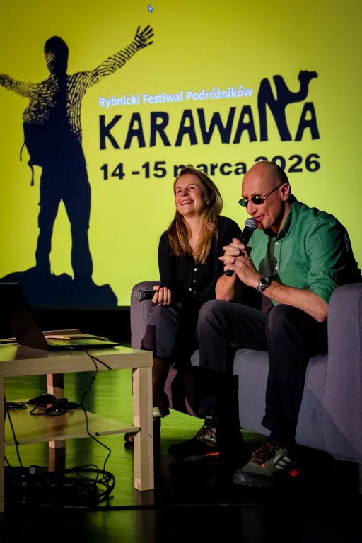  Kobieta i mężczyzna siedzą na kanapie na scenie. Przed nimi mały stolik z laptopem, za nimi wyświetlana rzutnikiem grafika “Karawana”. Kobieta uśmiecha się, a mężczyzna obok niej mówi do mikrofonu.