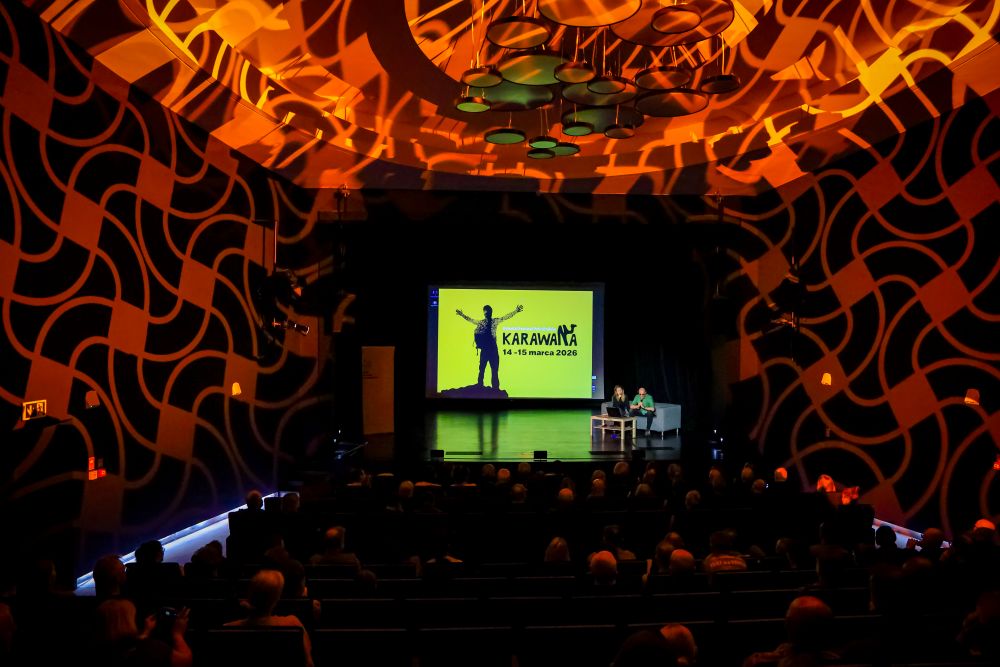 Scena, na której znajduje się kanapa, a na niej - kobieta i mężczyzna. Za nimi wyświetlana rzutnikiem grafika “Karawana”. Na ścianach oraz suficie wokół sceny widnieją różnorodne wzory. Przed sceną widać publiczność. 