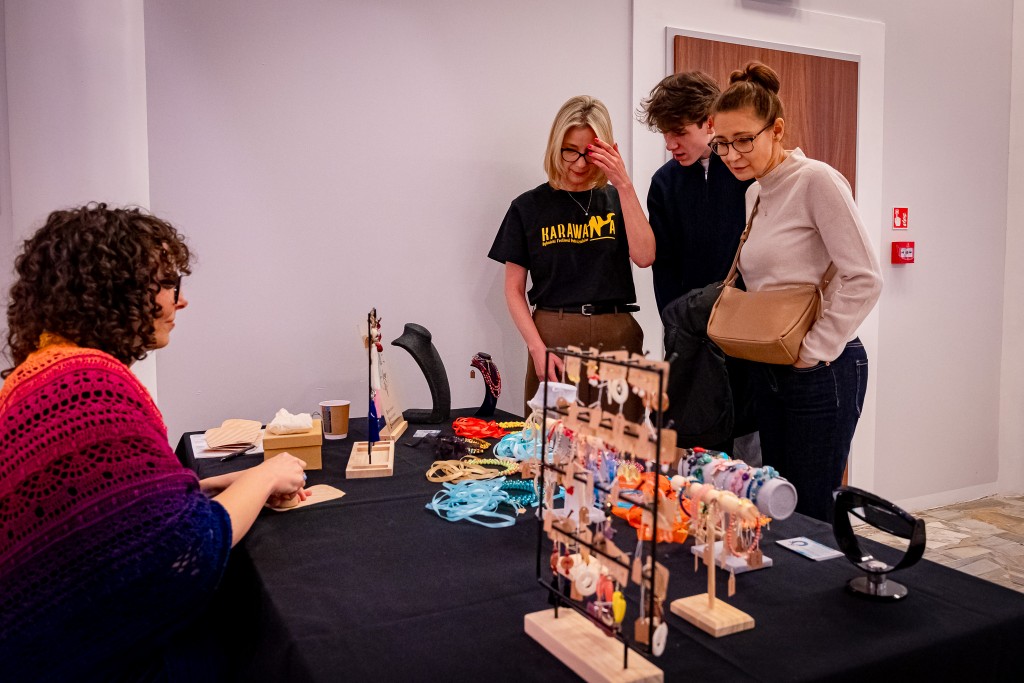 Dwie kobiety oraz mężczyzna stoją przy stoliku oraz przyglądają się biżuterii, która jest na nim wystawiona. Po drugiej stronie siedzi sprzedawczyni.