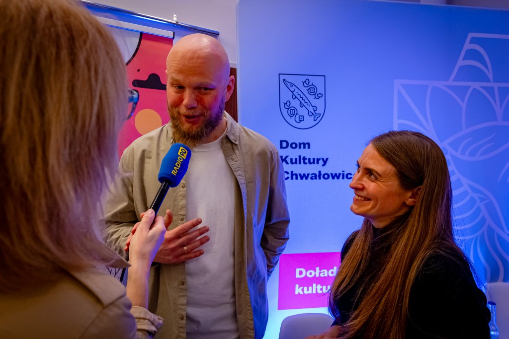 Mężczyzna udziela wywiadu kobiecie trzymającą mikrofon “Radio 90”, a obok niego stoi druga, uśmiechająca się kobieta.