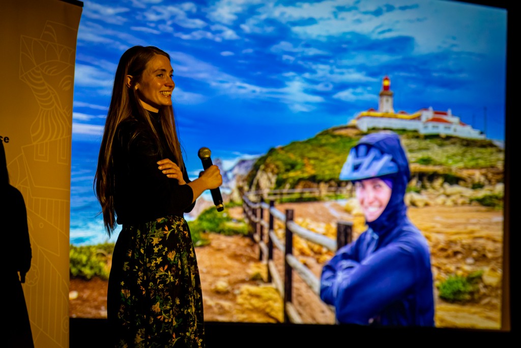 Kobieta uśmiecha się oraz trzyma mikrofon w ręce. W tle na rzutniku wyświetlane jest jej zdjęcie w stroju kolarskim na tle krajobrazu.