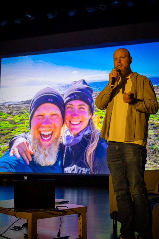 Mężczyzna mówi do mikrofonu, obok niego znajduje się stolik z laptopem.  W tle wyświetlane rzutnikiem jest zdjęcie jego z kobietą na tle krajobrazu - para uśmiecha się szeroko.