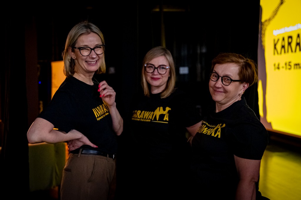 Trzy kobiety uśmiechają się do kamery. Mają na sobie koszulki z logiem “Karawana”, za nimi widoczna jest scena.