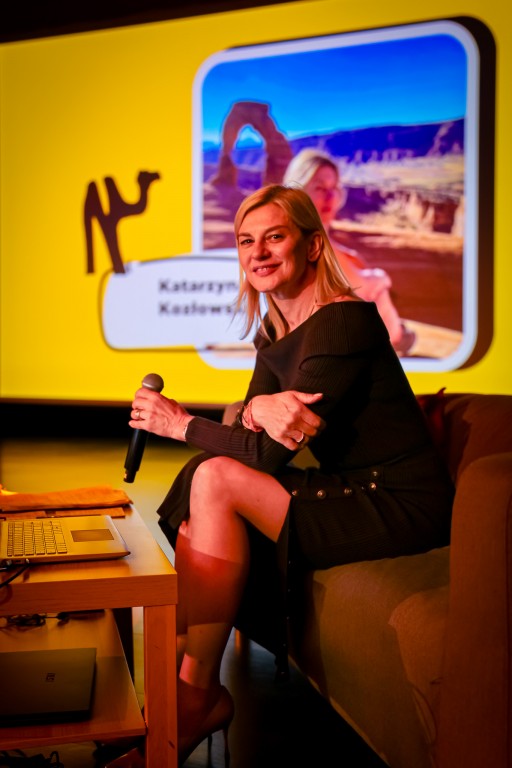 Kobieta siedzi na kanapie trzymając mikrofon i uśmiecha się do kamery.  W tle na rzutniku wyświetlane jest jej zdjęcie na tle krajobrazu.