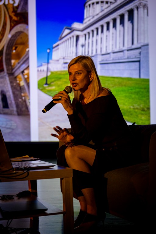 Kobieta siedzi na kanapie oraz mówi do mikrofonu. Przed nią znajduje się stolik z laptopem.