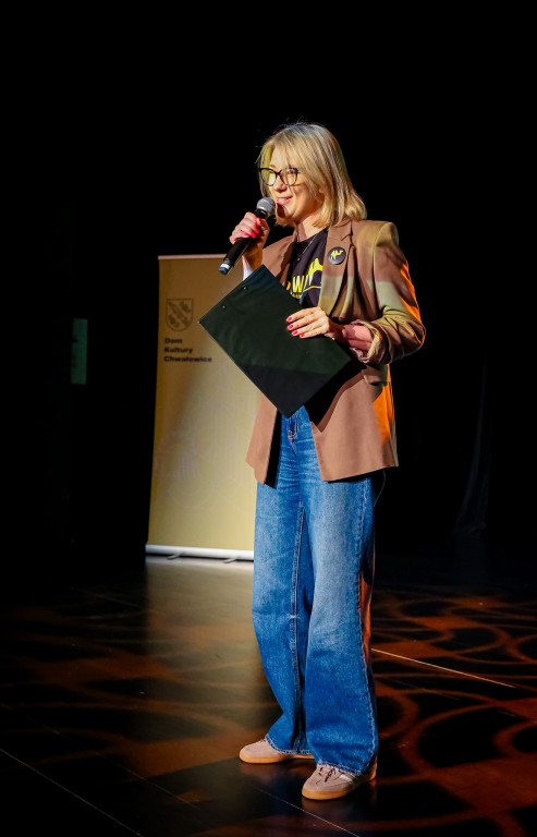 Kobieta stoi na scenie oraz mówi do mikrofonu, w drugiej ręce trzyma notes.