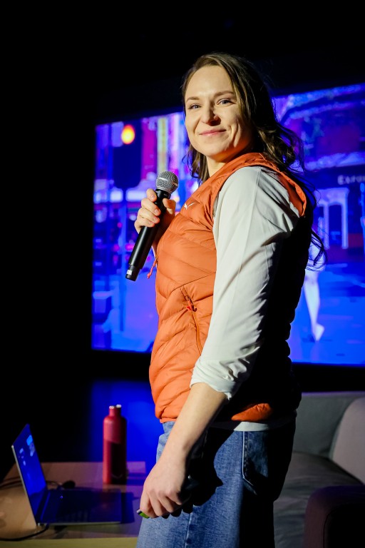 Kobieta trzyma w ręce mikrofon i uśmiecha się do kamery. W tle widoczna jest kanapa, stolik z laptopem oraz termosem, a za kobietą wyświetlany rzutnikiem obraz.