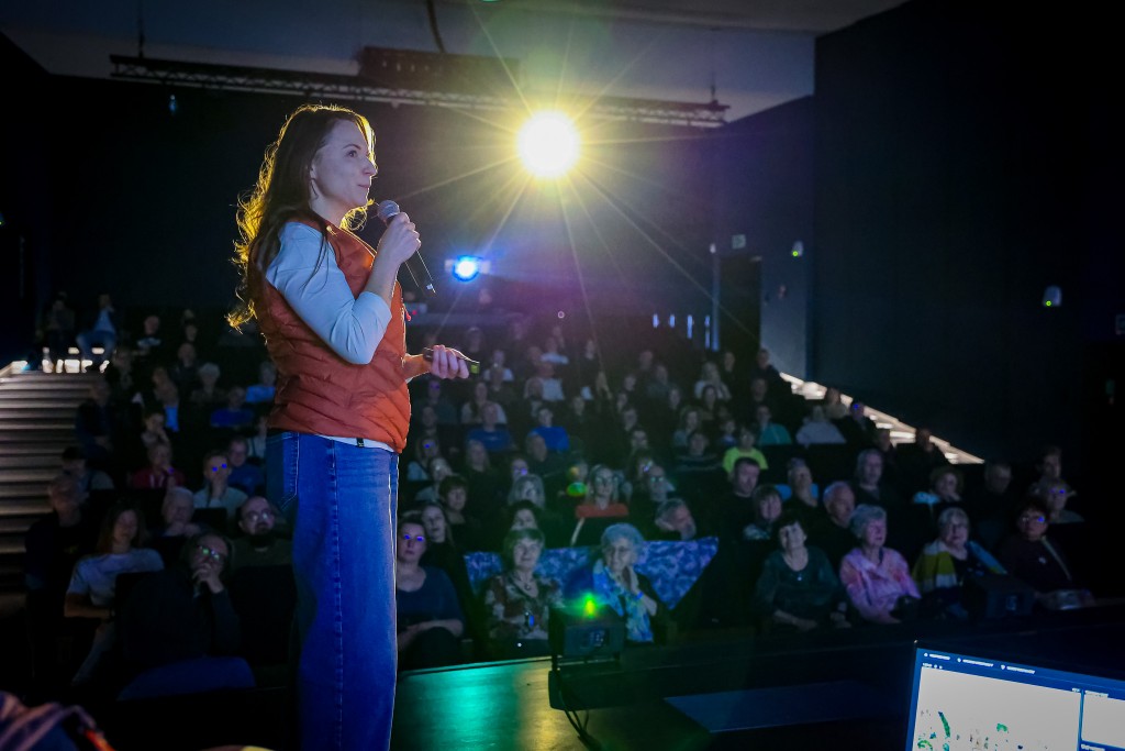 Kobieta mówi do mikrofonu oraz trzyma pilot od rzutnika. W tle widoczna publiczność.