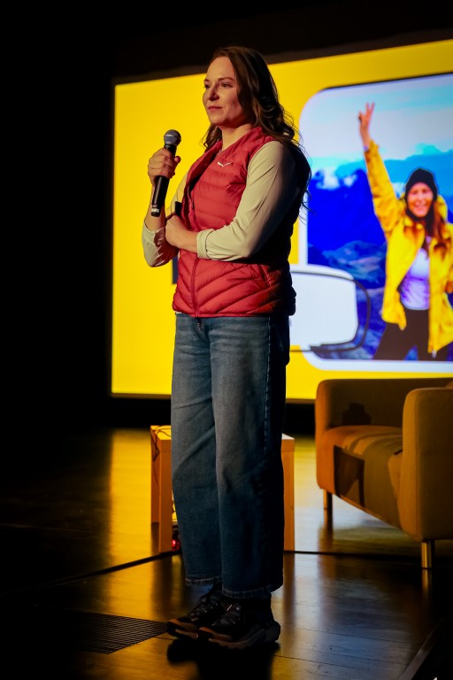 Kobieta trzyma w ręce mikrofon, za nią widoczna jest kanapa oraz stolik.  W tle wyświetlana rzutnikiem grafika ukazuje jej osobę w kurtce i czapce na tle krajobrazu.