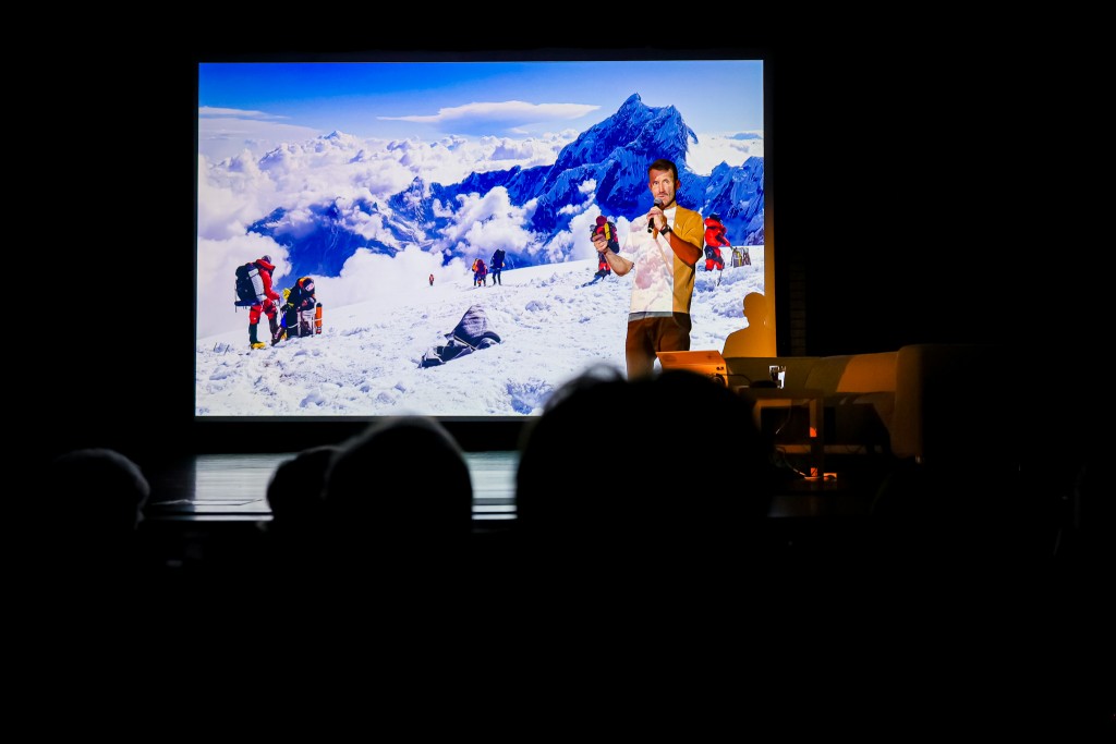 Mężczyzna mówi do mikrofonu oraz stoi na tle wyświetlanego rzutnikiem krajobrazu gór. Przed sceną jest częściowo widoczna publiczność.