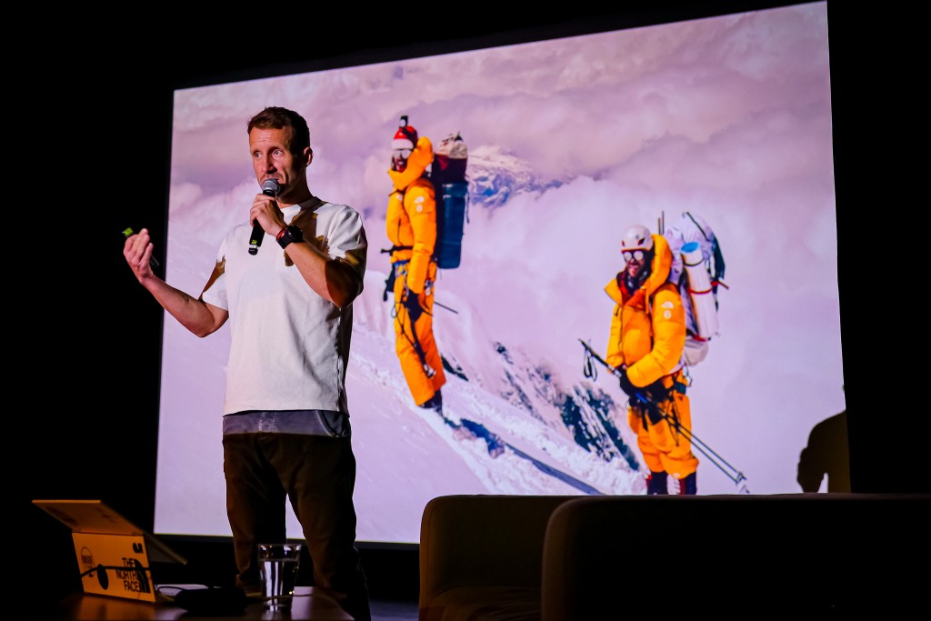 Mężczyzna mówi do mikrofonu, stoi na tle wyświetlanego rzutnikiem zdjęcia dwóch wspinaczy oraz krajobrazu gór.