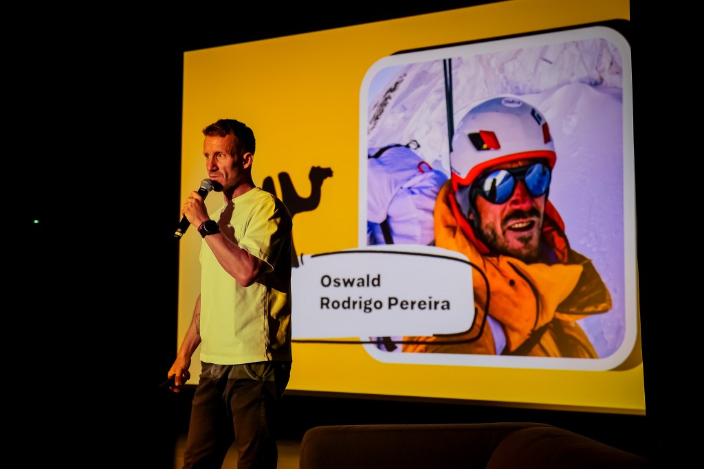 Mężczyzna mówi do mikrofonu, stoi na tle wyświetlanej rzutnikiem grafiki jego osoby w goglach, kasku oraz kurtce wspinaczkowej