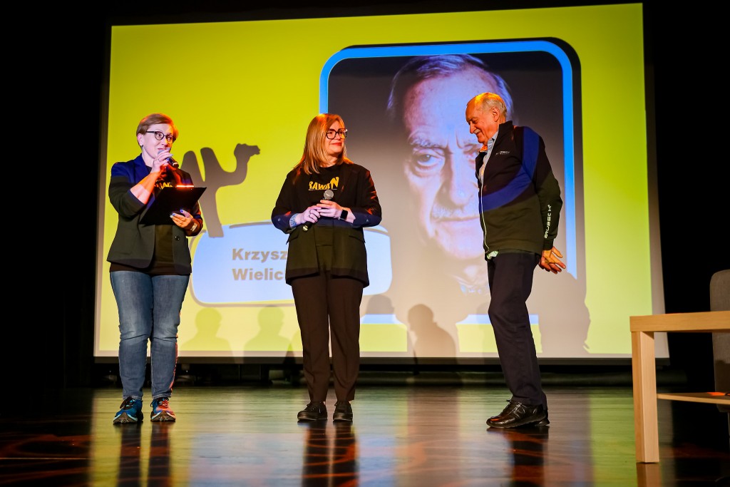 Dwie kobiety oraz mężczyzna stoją na scenie. Pierwsza kobieta mówi do mikrofonu i trzyma notes, druga trzyma mikrofon i uśmiecha się w stronę mężczyzny.  W tle na rzutniku wyświetlane jest jego zdjęcie.