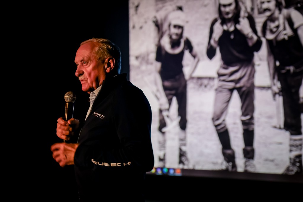 Mężczyzna mówi do mikrofonu, w tle na rzutniku wyświetlane jest stare zdjęcie trzech osób.