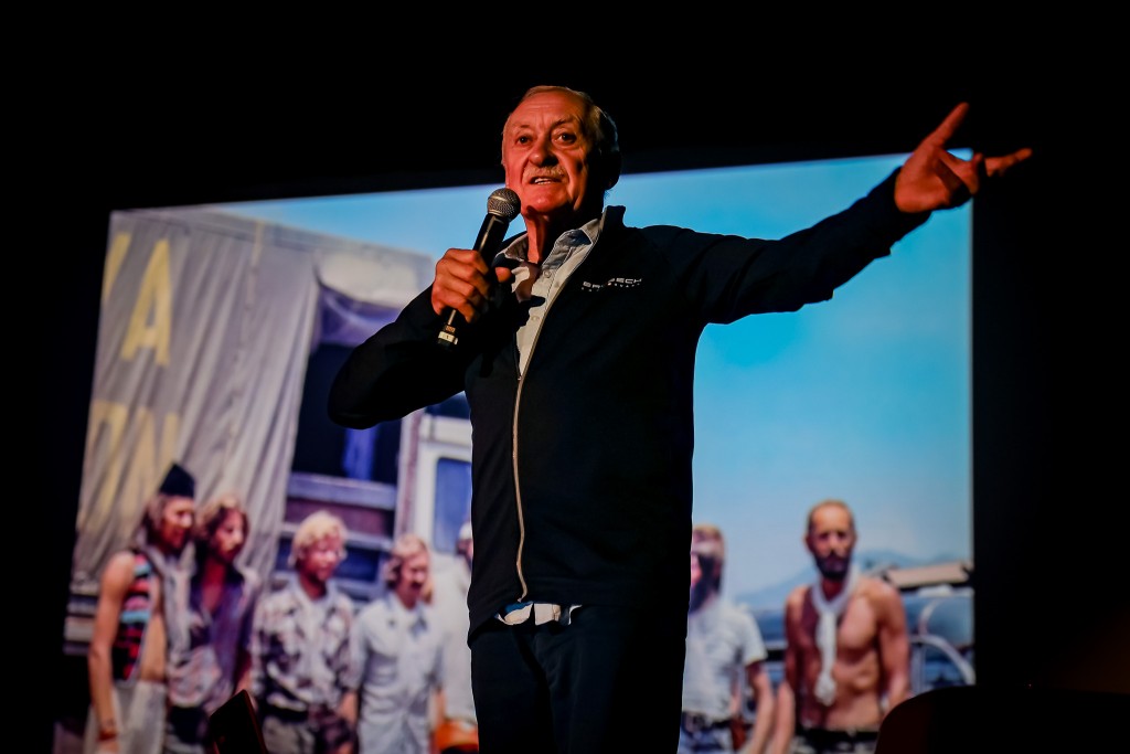 Mężczyzna mówi do mikrofonu, a za nim na rzutniku wyświetlane jest zdjęcie grupy mężczyzn