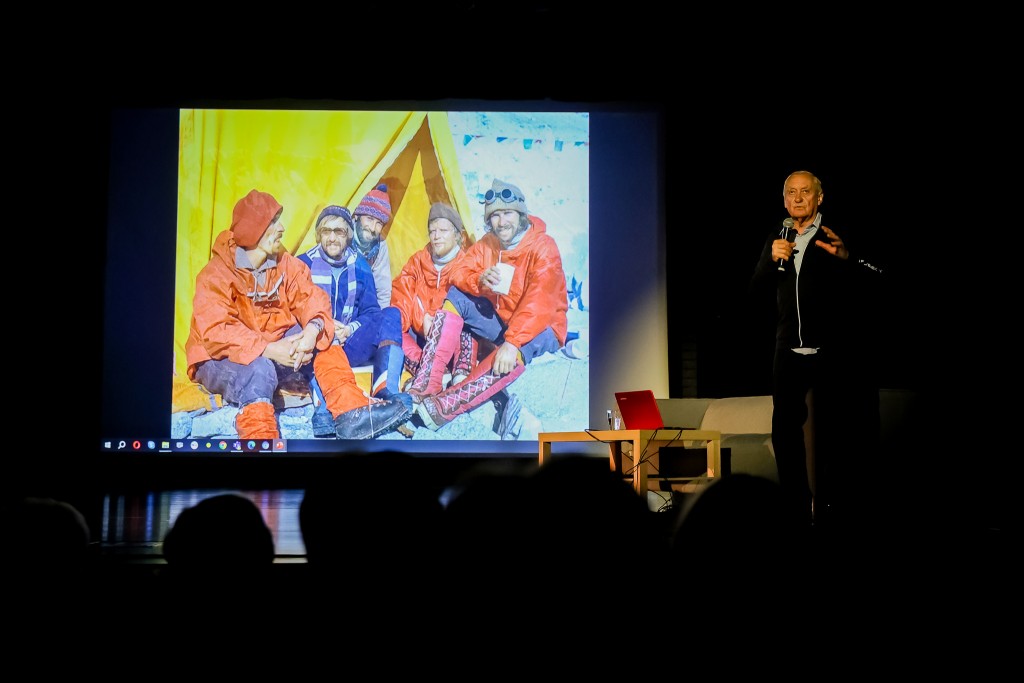Mężczyzna mówi do mikrofonu, obok niego znajduje się kanapa oraz stolik z laptopem. W tle wyświetlane rzutnikiem jest zdjęcie grupy wspinaczy siedzących przed namiotem.