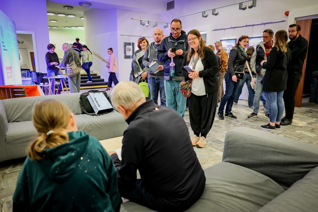 Dwie osoby siedzą na kanapie tyłem do obiektywu. Przed nimi grupa osób z książkami. 