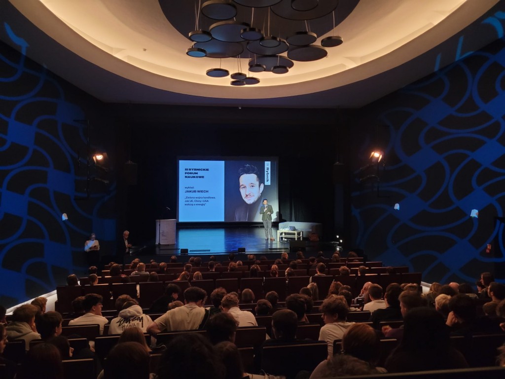 sytuacja sceniczna, mężczyzna na scenie mówi do publiczności, w tle prezentacja. Sala oświetlona na niebiesko