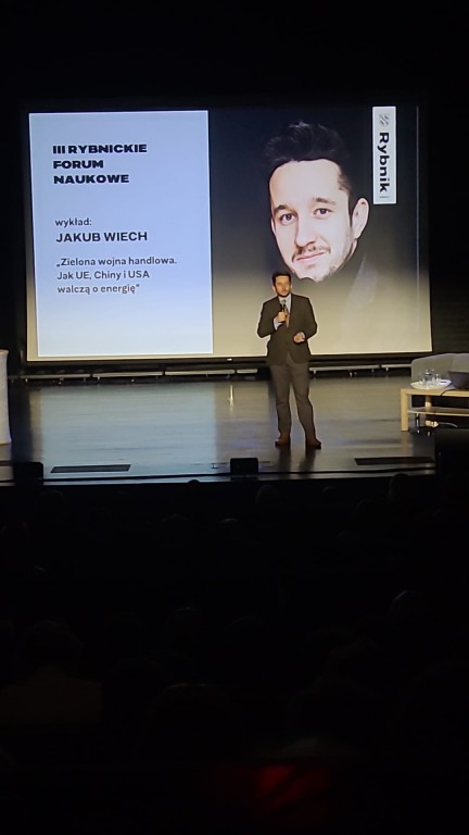 sytuacja sceniczna, mężczyzna na scenie mówi do publiczności, w tle prezentacja. Sala oświetlona na niebiesko