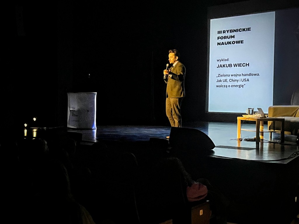 sytuacja sceniczna, mężczyzna na scenie mówi do publiczności, w tle prezentacja. Sala oświetlona na niebiesko
