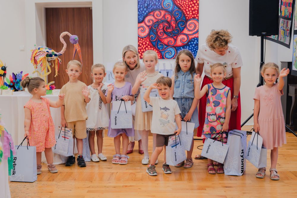 zdjęcie grupowe, małe dzieci z torebkami, część macha w kierunku osoby robiącej zdjęcia. Z tyłu kobieta.