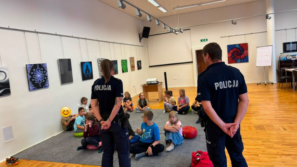 Zdjęcie grupowe, kilkoro dzieci siedzi na dywanie, przed nimi stoi Policjant i Policjantka. Twarz jednego dziecka zasłonięta emoji.
