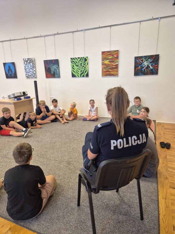 Grupa dzieci siedzi w okręgu na dywanie, na krześle w kręgu siedzi policjantka.