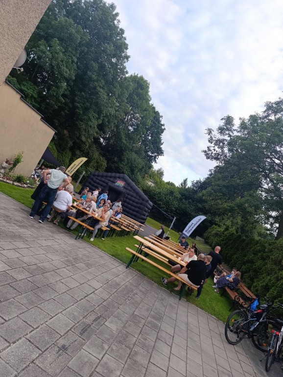 widok na 2 rzędy ław i stołów w plenerze, w tle dmuchany namiot, przy niektórych ławach siedzą już ludzie.
