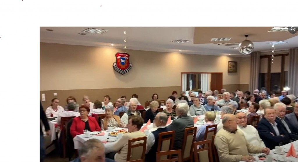 grupa osób przy 3 długich bankietowych stołach siedzi na krzesłach.