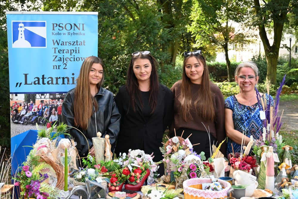 4 kobiety stoją za stołem zastawionym rękodziełem (figurki, kompozycje kwiatowe), w tle baner PSONI koło w Rybniku.