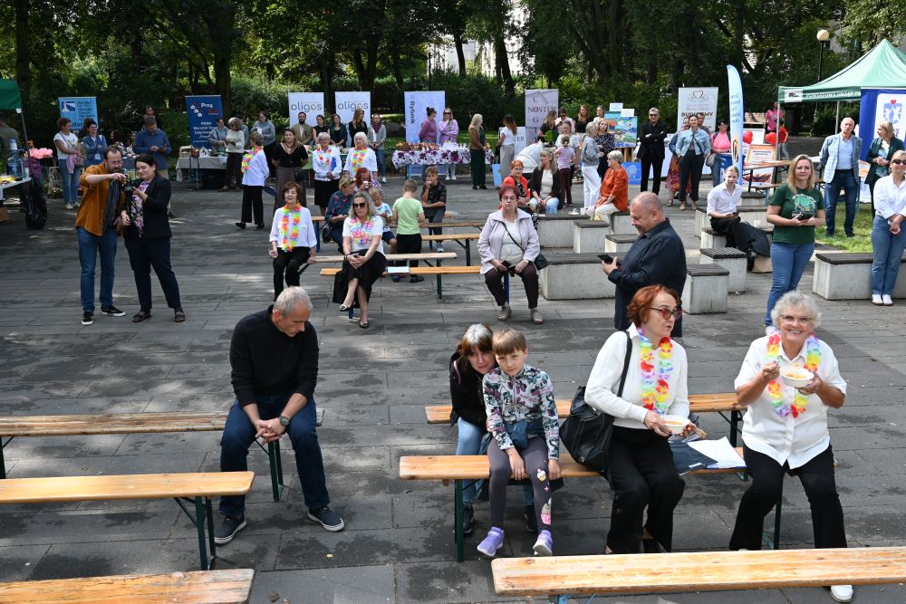 Plener, widok na plac gdzie rozłożono piknikowe ławki, na niektórych siedzą ludzie, wokół placu rozstawiono stoiska, niektóre zadaszają namioty.