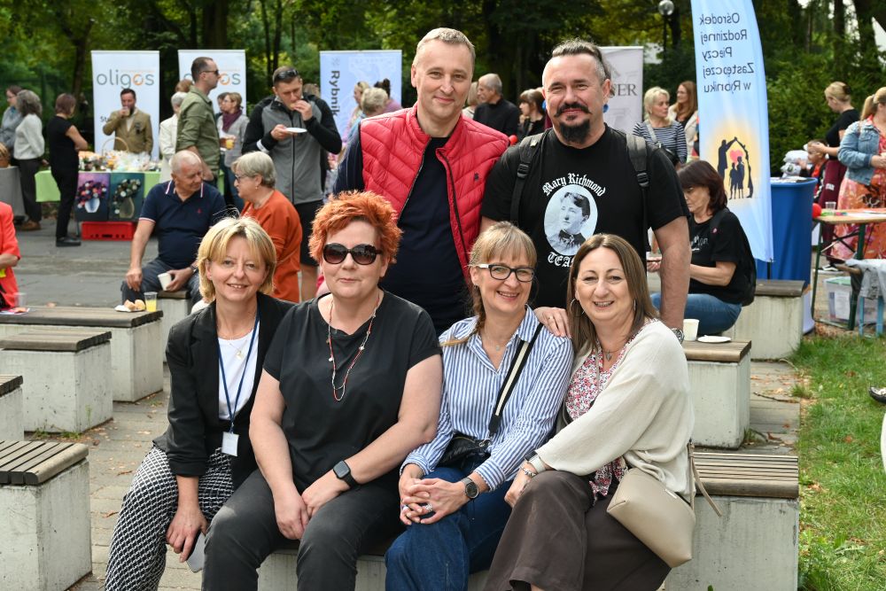 Grupa osób, plener. Cztery osoby siedzą na ławce, w tle stoi dwóch mężczyzn. Pozują do zdjęcia.