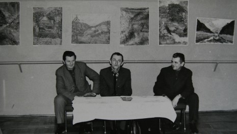 Trzech mężczyzn siedzących przy stole, za nimi na ścianie zdjęcia