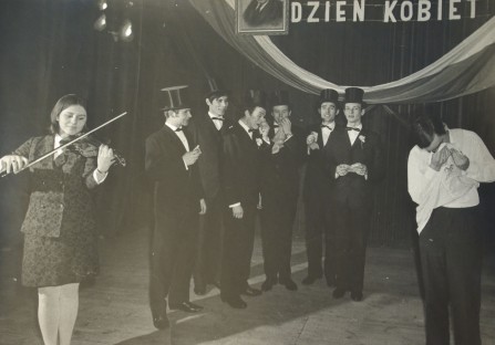Występ, kobieta gra na skrzypcach, siedmiu mężczyzn stoi elegancko ubranych, jeden z mężczyzn przeciera twarz chusteczką
